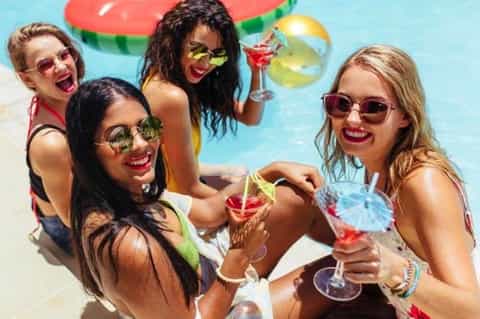 Group of four women by pool wearing sunglasses holding colorful tropical cocktails and smiling