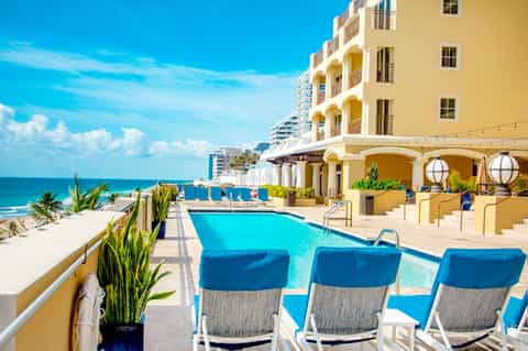 Beachfront resort with yellow building, blue pool, ocean view, and palm trees under clear sky