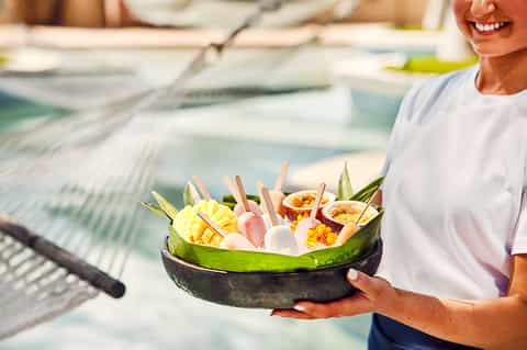 Staff member holding coconut bowl of tropical appetizers by resort pool in daylight