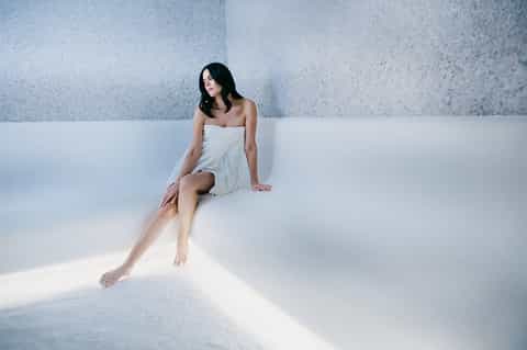 Woman in white wrap seated against textured wall with dramatic natural sunlight