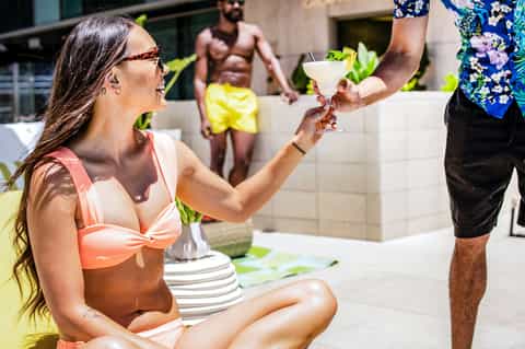Woman in bikini receiving cocktail at poolside party with guests