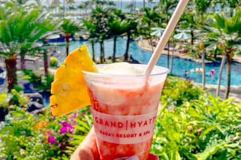 Tropical cocktail with pineapple garnish overlooking Grand Hyatt Kauai Resort pool and gardens