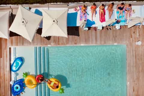 Overhead view of pool with people sunbathing, float toys, umbrellas, and wooden deck beside water