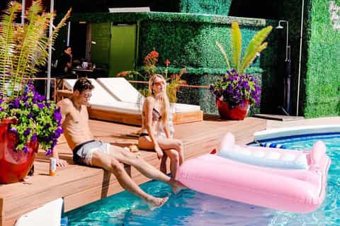 Resort pool area with couple relaxing on deck near pink inflatables, green living wall backdrop