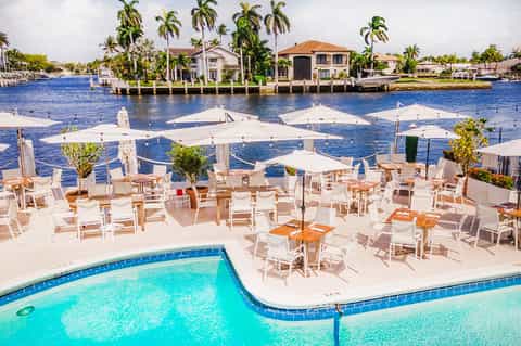 Waterfront resort pool with white umbrellas, lounge chairs, and waterfront houses in the background