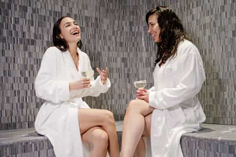 Two women in white robes enjoying beverages in a spa relaxation area with textured tile walls