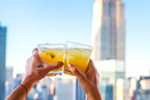 Two hands toasting with glasses of amber cocktails against blurred city skyline backdrop