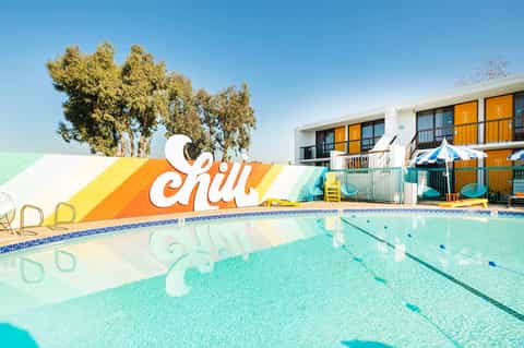 Retro-style outdoor swimming pool with 'Chill' signage, yellow umbrellas, and mid-century building