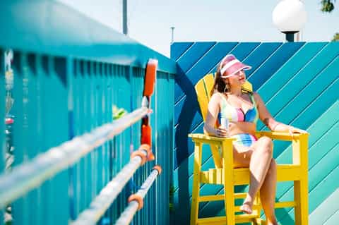 Woman in colorful bikini sitting on yellow lifeguard chair by turquoise pool fence with buildings