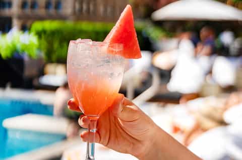 Guest holding watermelon-garnished cocktail by poolside with blurred resort background