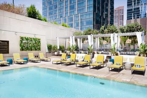 Modern urban pool deck with yellow lounge chairs, white pergolas, and city building backdrop