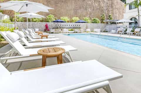 Resort pool deck with white lounge chairs, umbrellas, stone walls, and manicured landscaping