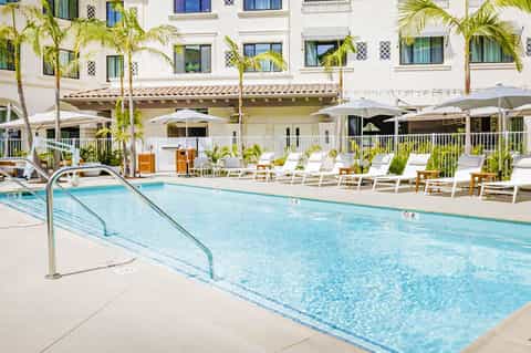 Resort swimming pool with loungers, umbrellas, palm trees, and white Mediterranean-style building