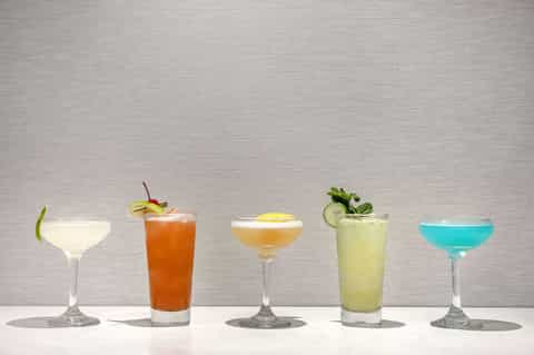 Five tropical cocktails in various colors displayed on white surface with garnishes