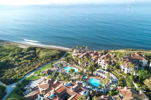 Aerial view of beachfront resort with multiple pools, palm trees, Mediterranean architecture, and ocean