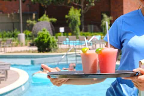 Server in blue uniform carrying tray with colorful cocktails by resort pool