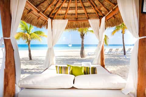 Beachfront cabana bed with white pillows and flowing curtains overlooking ocean and palm trees