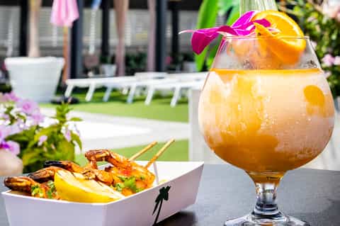 Tropical cocktail with orange slice and flower garnish, grilled shrimp appetizer, poolside resort background