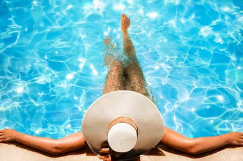 Person floating in clear blue pool wearing white sun hat, viewed from above