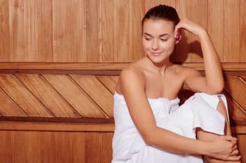 Woman in white towel relaxing against wooden sauna wall paneling