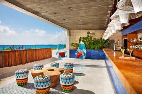 Modern beachfront bar lounge with woven pendant lights, colorful seating, ocean views, and wooden accents