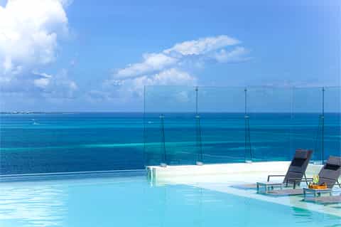 Infinity pool with glass railing overlooking vibrant turquoise ocean and white cloud sky