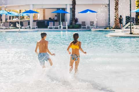 Children running into resort pool with blue umbrellas and palm trees in background