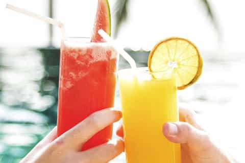 Two tropical drinks held by hands overlooking a swimming pool and beach loungers