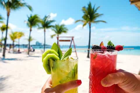 Tropical beach cocktails with fresh fruit garnishes held by hands with palm trees and beach in background