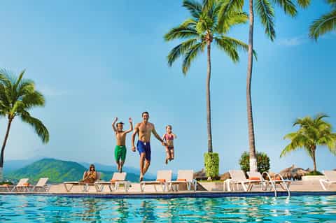 Family jumping into tropical pool with palm trees, mountains, and lounge chairs