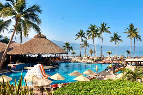 Tropical beachfront resort pool with thatched-roof bar, palm trees, umbrellas, and ocean view in background