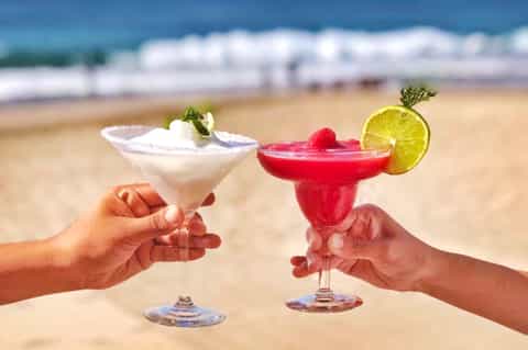 Two colorful tropical cocktails held on beach with ocean waves in background