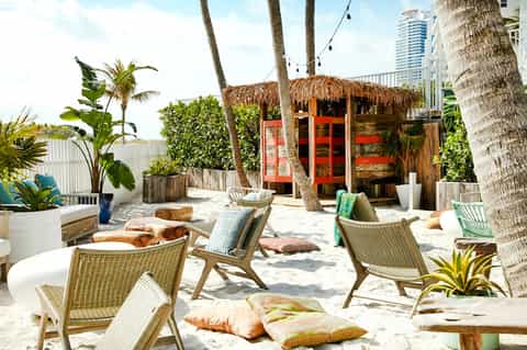 Beach cabana lounge with thatched roof, palm trees, wooden chairs, and colorful pillows on white sand