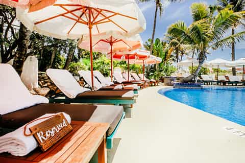 Poolside lounge area with white cushioned chairs under red and white umbrellas among palm trees