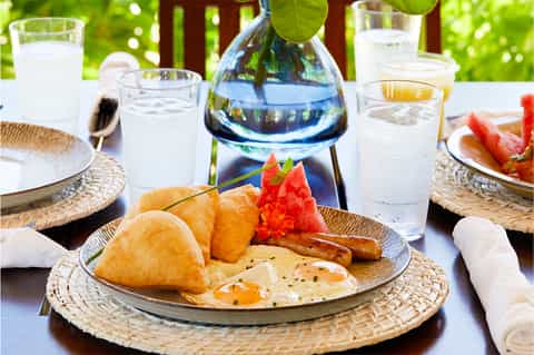 Outdoor dining table set with tropical breakfast including pastries, fruit, eggs, beverages, and ocean view backdrop
