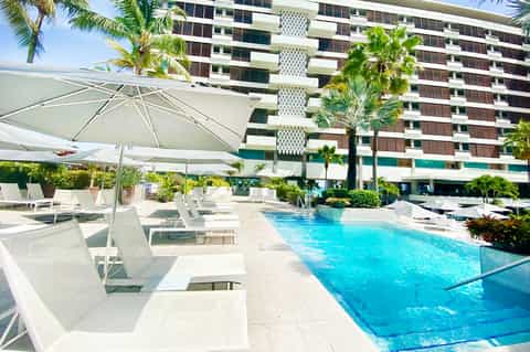 Resort pool with white umbrellas, loungers, high-rise hotel building, and palm trees under clear blue sky