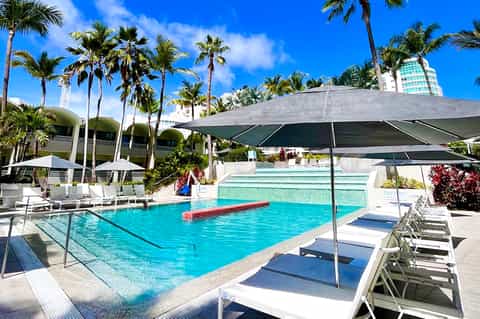 Resort pool area with turquoise water, palm trees, lounge chairs, and umbrellas under blue sky