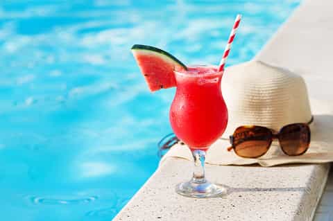 Red tropical cocktail with watermelon slice and striped straw poolside with sunglasses and sun hat