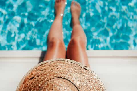 Person's legs dangling over clear turquoise pool water while wearing straw hat