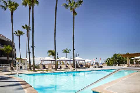 Outdoor resort pool surrounded by tall palm trees with white cabanas and umbrellas under clear blue sky