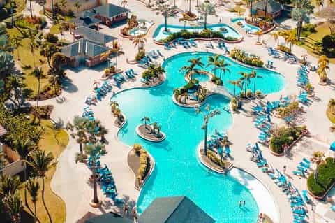 Aerial view of large resort pool complex with turquoise water, palm trees, lounge chairs, and sandy areas