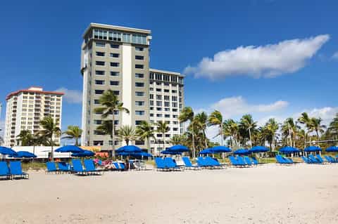 Beachfront hotel tower with blue umbrellas and lounge chairs on sandy beach, palm trees, and clear sky