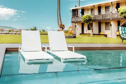 White lounge chairs partially submerged in turquoise pool with coastal resort building and lawn