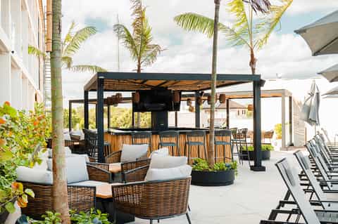 Modern outdoor bar with woven seating, black pergola, and palm trees alongside pool area