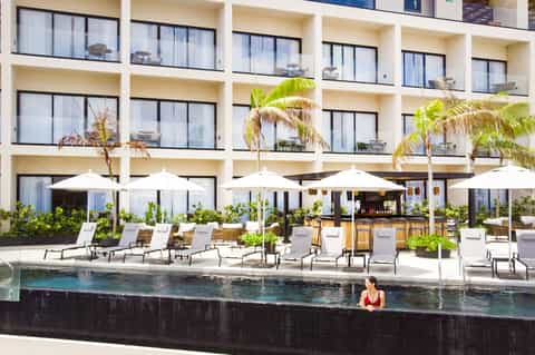 Modern beachfront hotel with white umbrellas, loungers, palm trees, and woman swimming