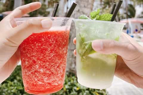 Two colorful frozen slushie drinks in plastic cups with straws held at a beachfront location