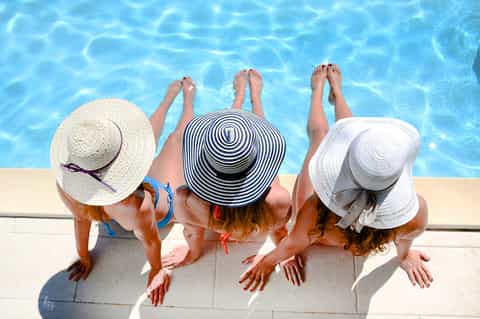 Three women in sun hats relaxing poolside with feet dangling in clear blue water