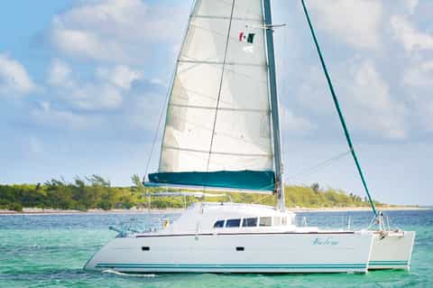 White catamaran sailboat with blue sail on turquoise water near tropical beach with mangroves