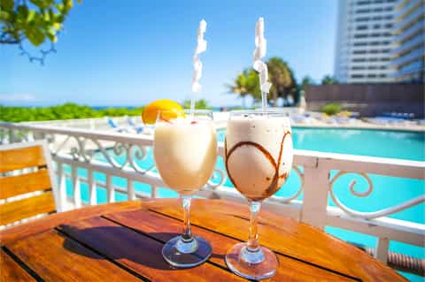 Two tropical frozen cocktails with straws by pool overlooking ocean and palm trees