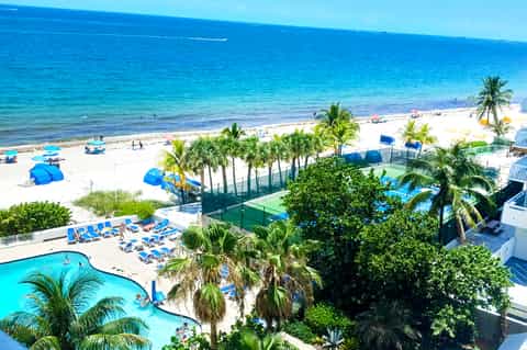 Beachfront resort with blue lounge chairs, pool, sports facilities, palm trees, and ocean view